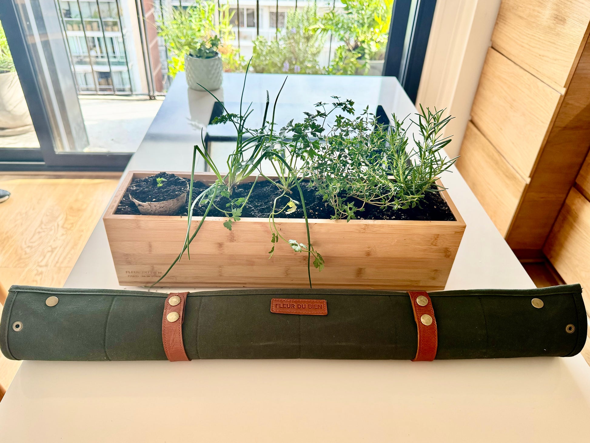 Fleur Du Bien 30x30 inch green indoor repotting mat folded next to the herb kit with planter, displayed in a luxury French-inspired minimalist kitchen with oak tables and Silestone countertops. Perfect for indoor gardening, potting herbs, transplanting, watering, and mess-free plant care. Stylish, eco-friendly, and ideal for elegant home décor and DIY gardening projects.