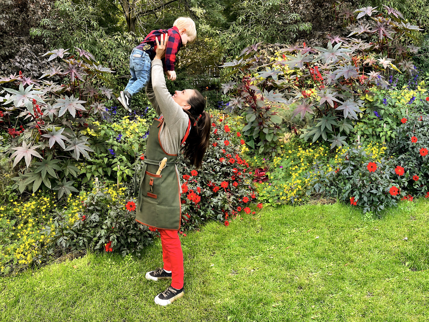 Mom wearing Fleur Du Bien apron holding her 2-year-old child up in the air in a stunning garden with a vibrant flower border, both smiling after planting herbs, showcasing family gardening fun and eco-friendly lifestyle