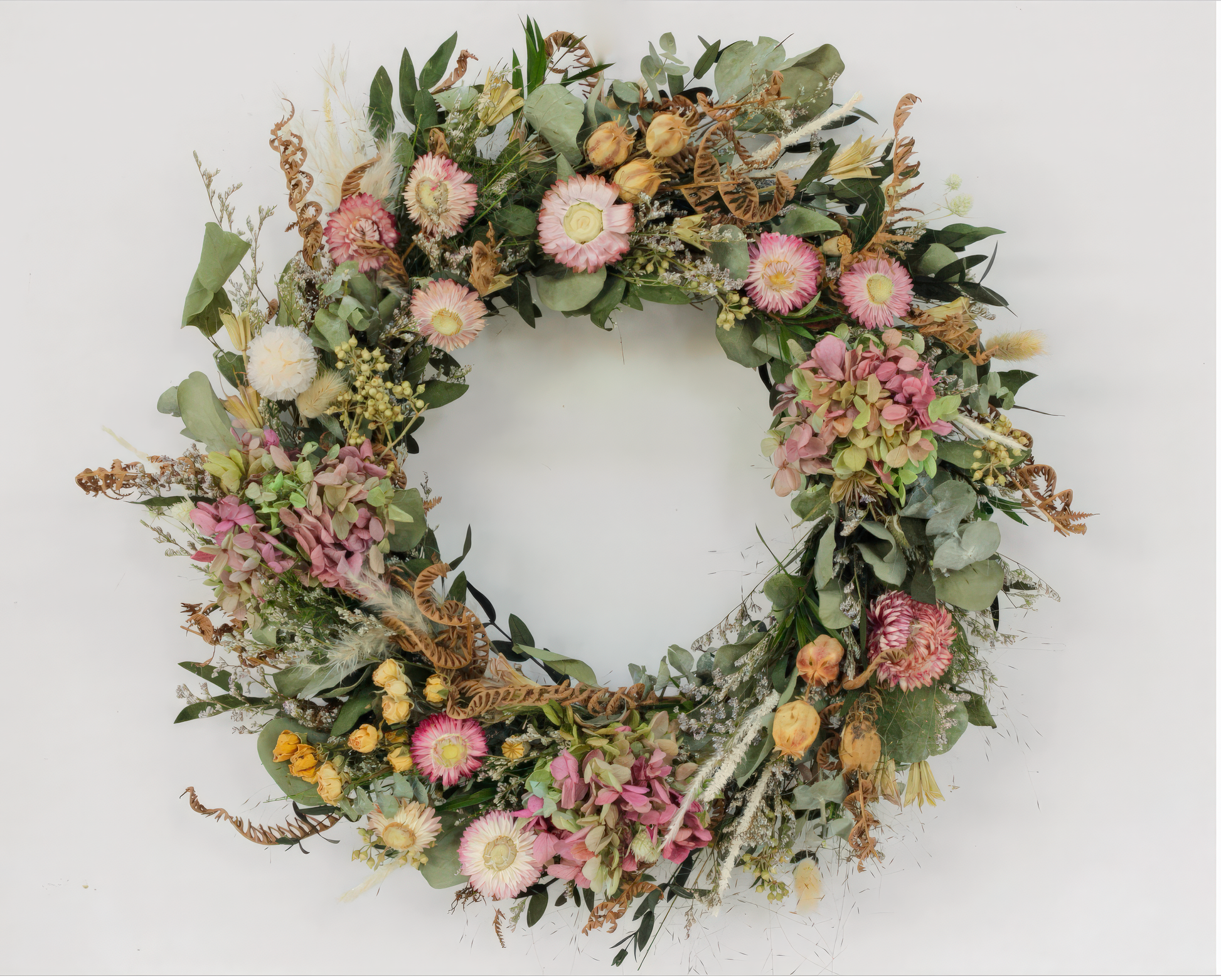 Fleur Du Bien Blush Blooms 16-inch dried flower wreath hanging on front door, natural hydrangea, chrysanthemum, roses, rabbit tails, eucalyptus. A round dried flower wreath featuring a variety of flowers including pink hydrangea, eucalyptus leaves, and other dried botanicals, arranged in a circle.