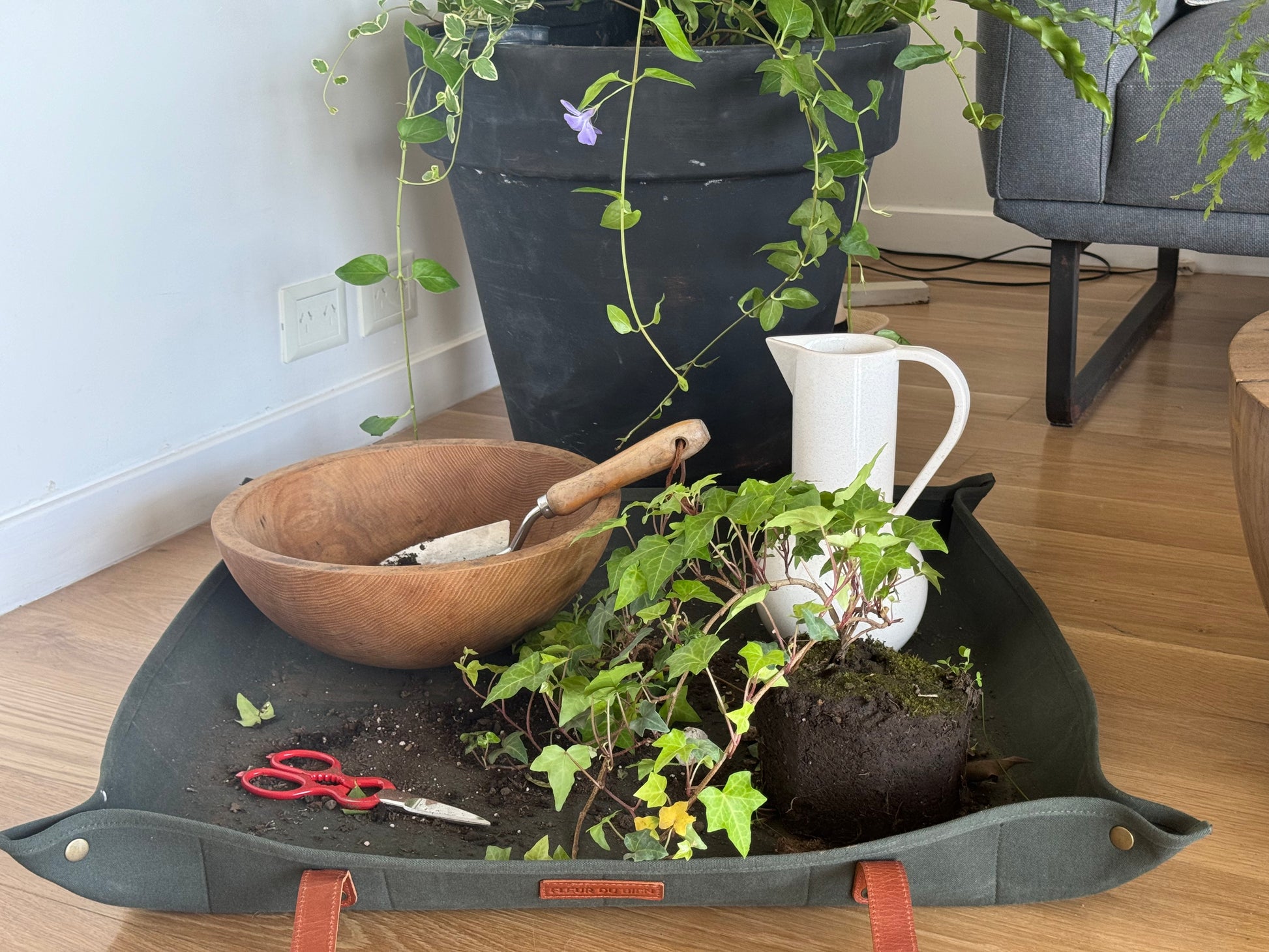 Fleur Du Bien 30x30 inch green indoor repotting mat with leather trims, featuring Vincas plants placed under a Ficus Rubber Tree pot to create a beautiful base. Includes Fleur Du Bien garden tools nearby, perfect for planting, transplanting, watering, and other indoor gardening projects. Eco-friendly, durable, and mess-free solution for all your herb and plant care needs