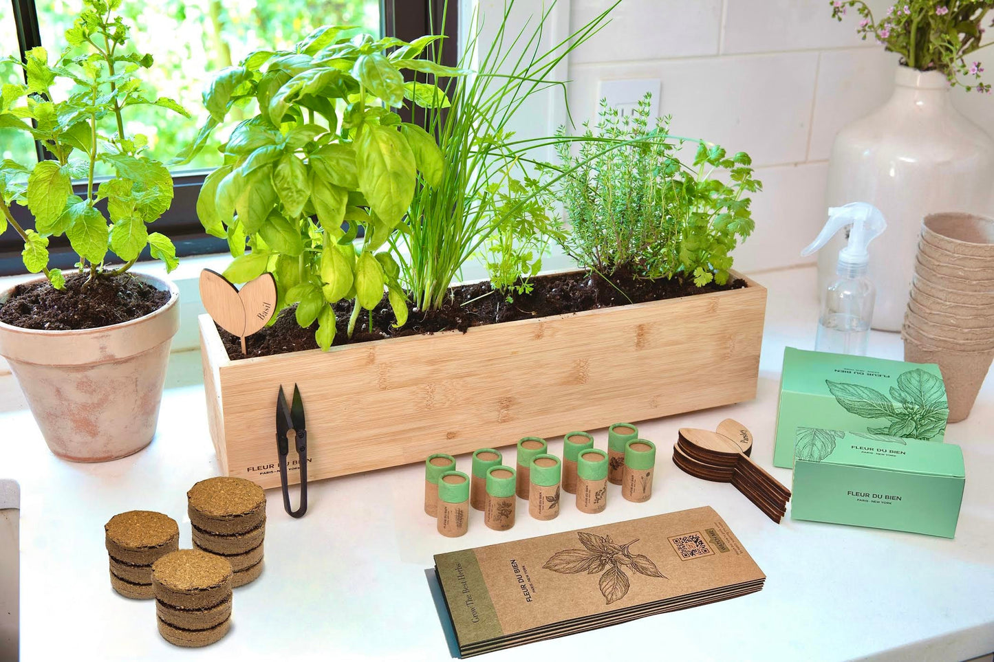 Fleur Du Bien Indoor Herb Garden Starter Kit with planter, 6 herb seeds, soil discs, and plant markers. An indoor herb garden kit setup including biodegradable growing pots, soil discs, plant markers, seed packets, and a wooden herb planter, displayed on a table.