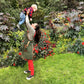 Mom wearing Fleur Du Bien apron holding her 2-year-old child up in the air in a stunning garden with a vibrant flower border, both smiling after planting herbs, showcasing family gardening fun and eco-friendly lifestyle